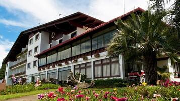 Guests can enjoy a tranquil garden setting with a variety of flowering plants and a palm tree, surrounded by a traditional hotel with large windows and outdoor seating areas.