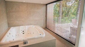 A modern bathroom with a freestanding bathtub, a glass shower enclosure, and a view of greenery through the window blinds.