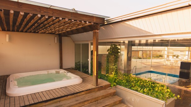 A hot tub under a wooden pergola with a view of a swimming pool and outdoor furniture.