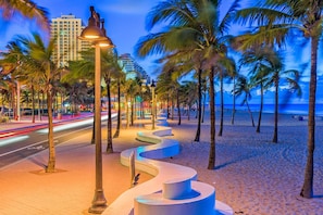 A beachside promenade with palm trees, street lamps, and a road with light trails.