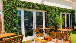 An outdoor patio with wooden tables and chairs, a breakfast spread, and a lush green wall with flowers.
