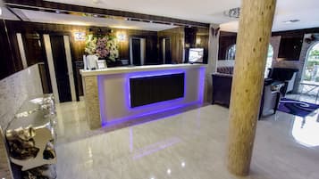 A hotel reception area with a modern design, featuring a marble floor, a reception desk with a flat-screen TV, and a large mirror above the desk.