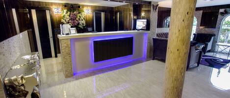 Zona de recepción de hotel con diseño moderno, que presenta un suelo de mármol, un mostrador de recepción con televisión de pantalla plana y un espejo grande sobre el mostrador.