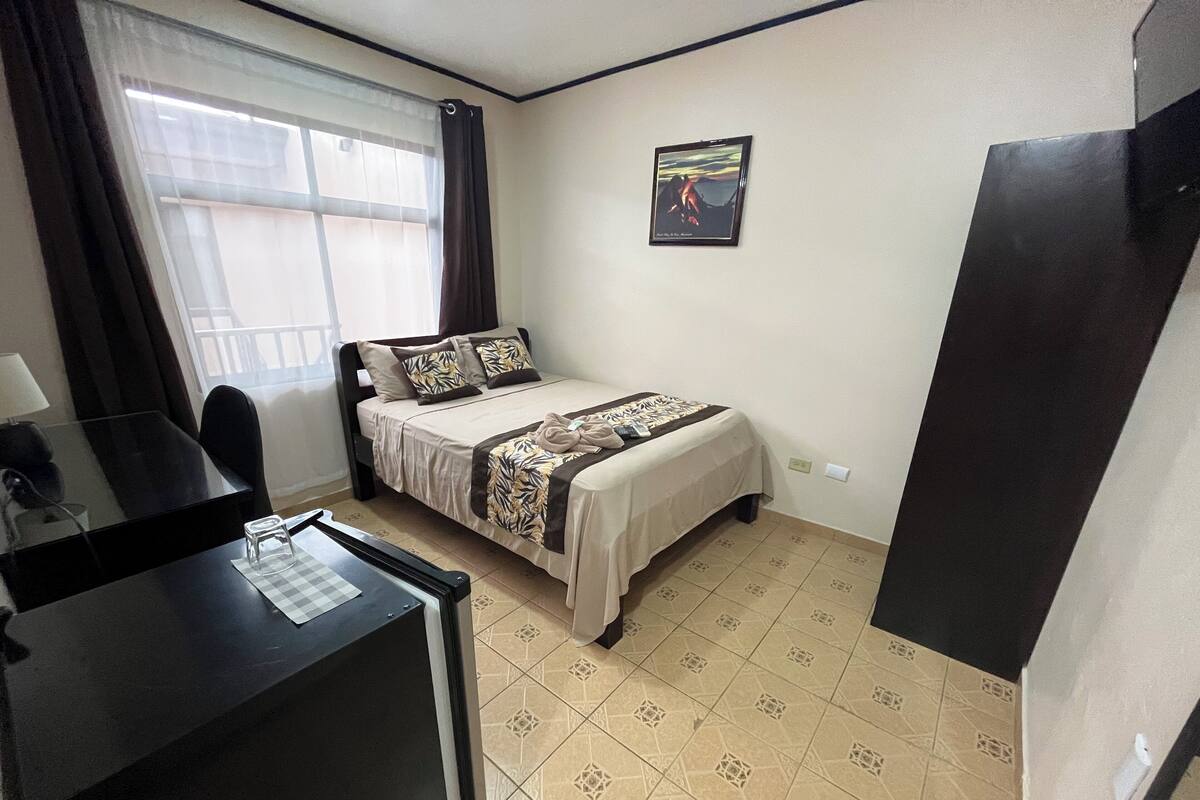 A small, tidy room with a bed, a desk, and a chair. There is a window with curtains, a framed picture on the wall, and a tiled floor.