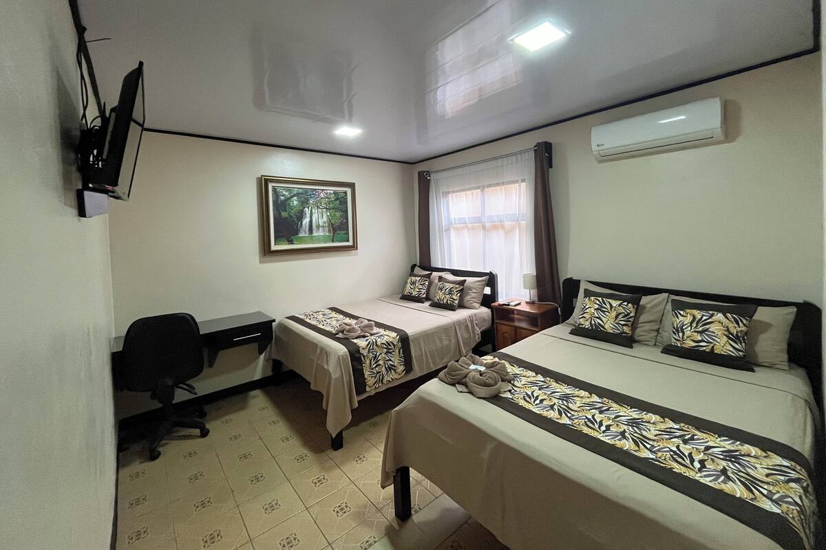 A hotel room with two beds, a desk, a chair, a TV, a painting, and an air conditioner.