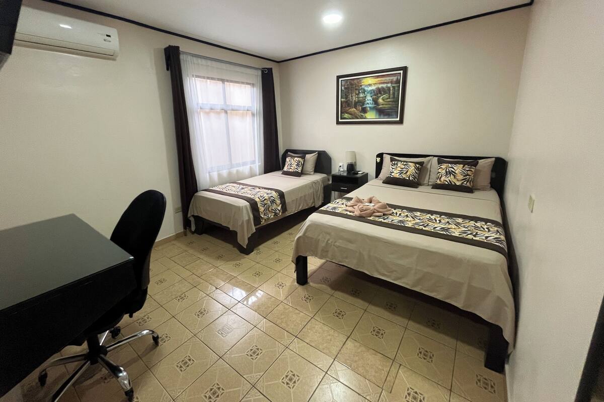 A hotel room with two single beds, a desk, a chair, a window with curtains, an air conditioning unit, and a painting on the wall.