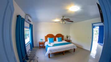 Un dormitorio con cama, ventilador de techo, equipo de aire acondicionado, mesita de noche, lámpara y ventana con cortinas.