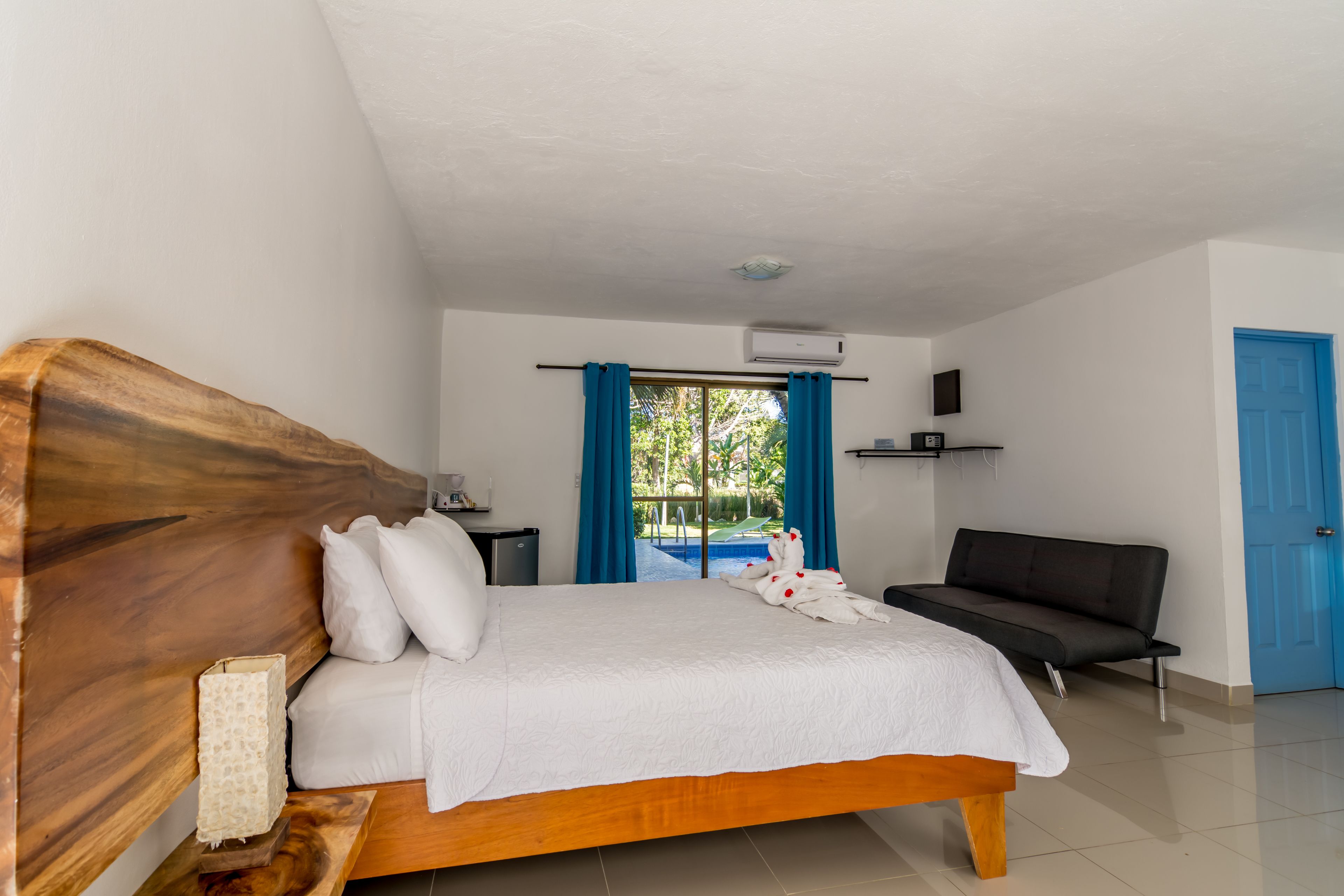 Un dormitorio con cabecera de madera, cama con sábanas blancas, sofá negro y vista a una piscina a través de una ventana con cortinas azules.