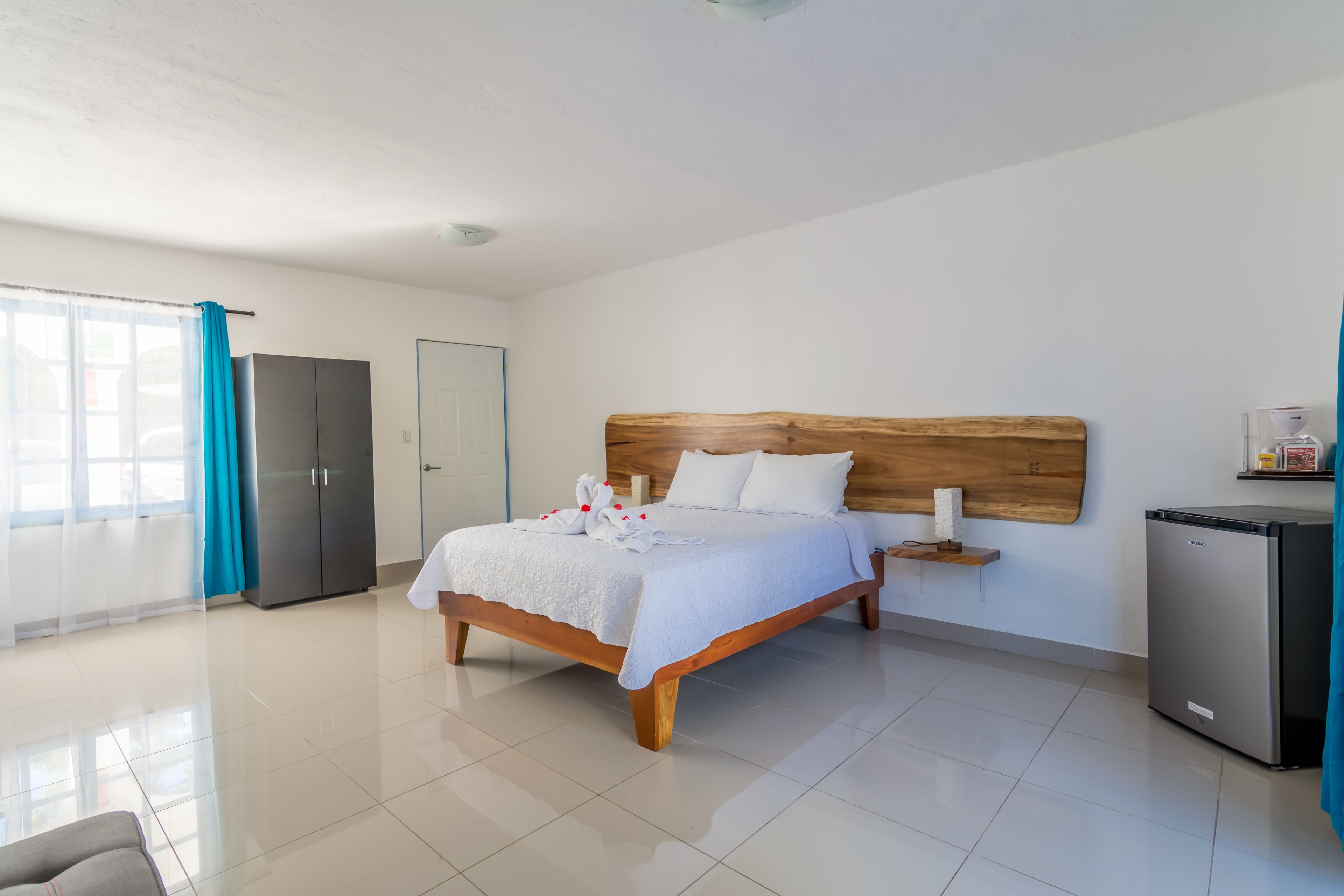 A modern hotel room with a large bed, a wooden headboard, a small bedside table, a refrigerator, and a bathroom with a shower and toilet.