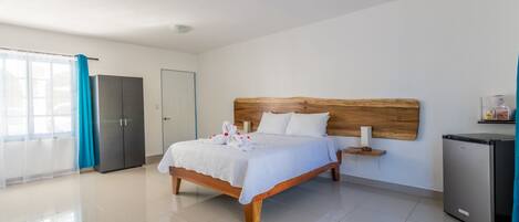 A modern hotel room with a large bed, a wooden headboard, a small bedside table, a refrigerator, and a bathroom with a shower and toilet.