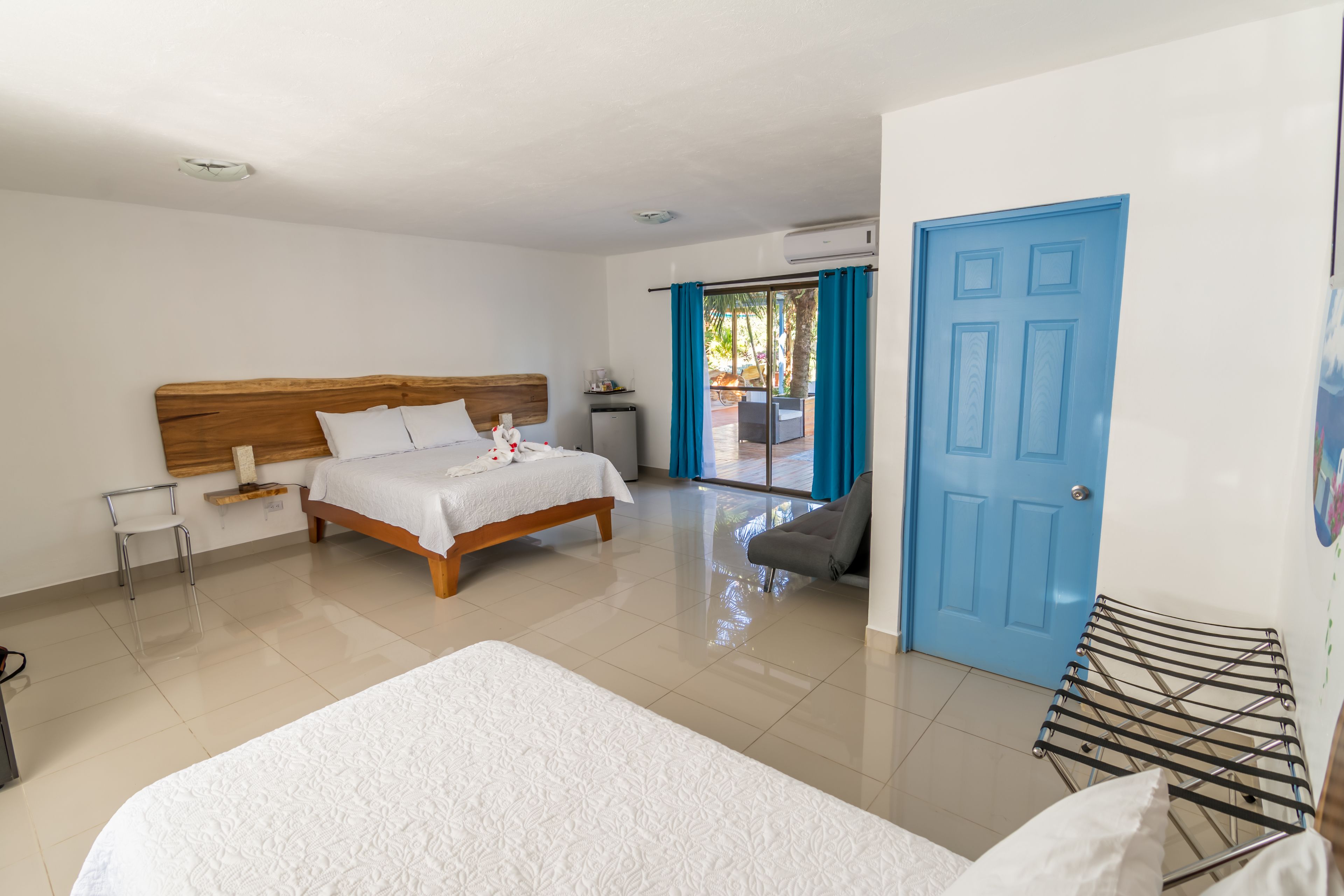 Une chambre d’hôtel avec un lit en bois, un deuxième lit, une petite table, une chaise, une porte bleue et une vue sur l’extérieur.