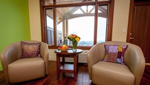 Two leather armchairs with patterned cushions, a small wooden table with a vase of flowers and a fruit bowl, and a large window with a view of a building and trees.