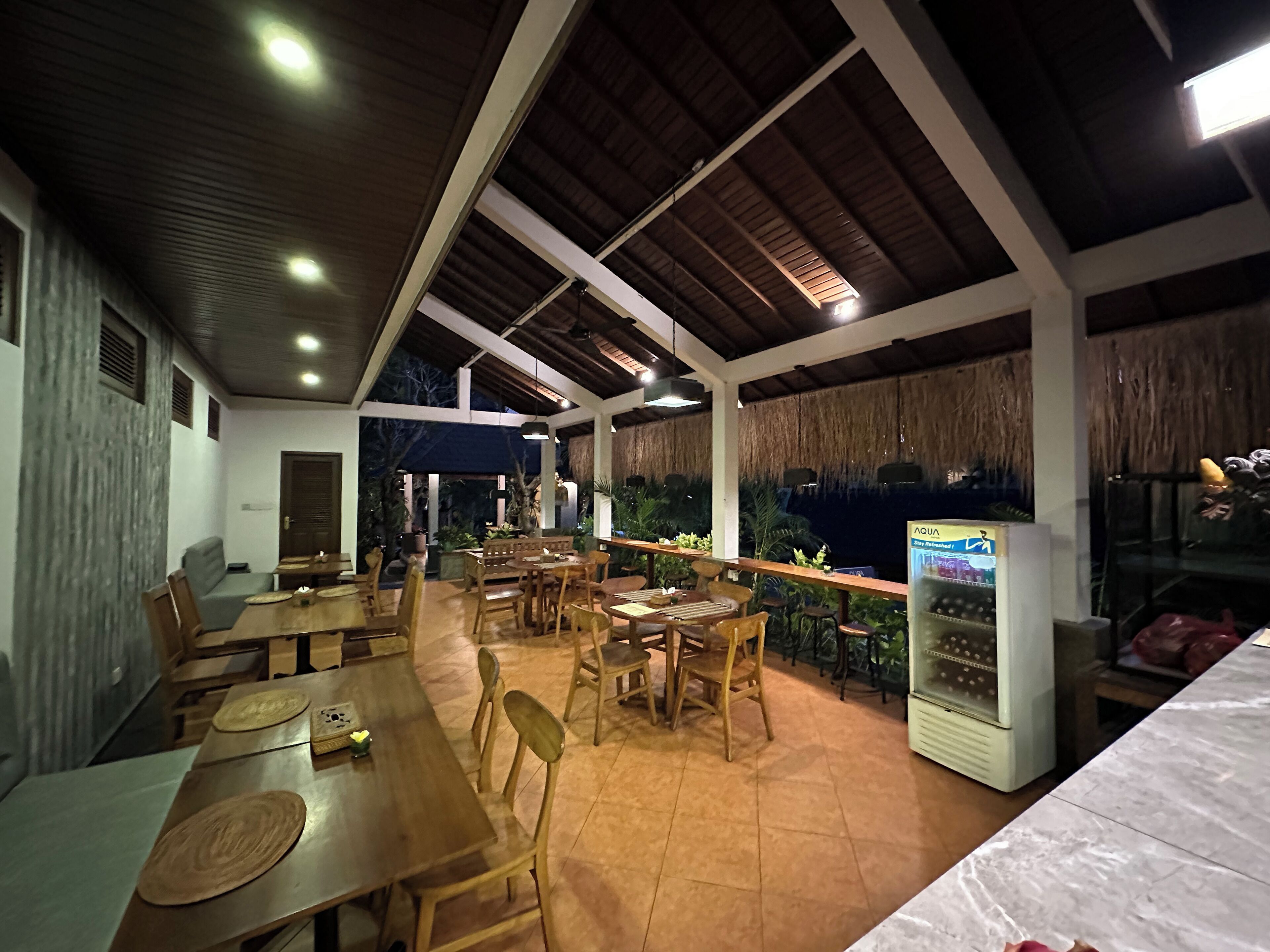 Un espace extérieur abrité, aménagé pour les repas, avec des tables et des chaises en bois, un mur recouvert de chaume et un élément décoratif aquatique.