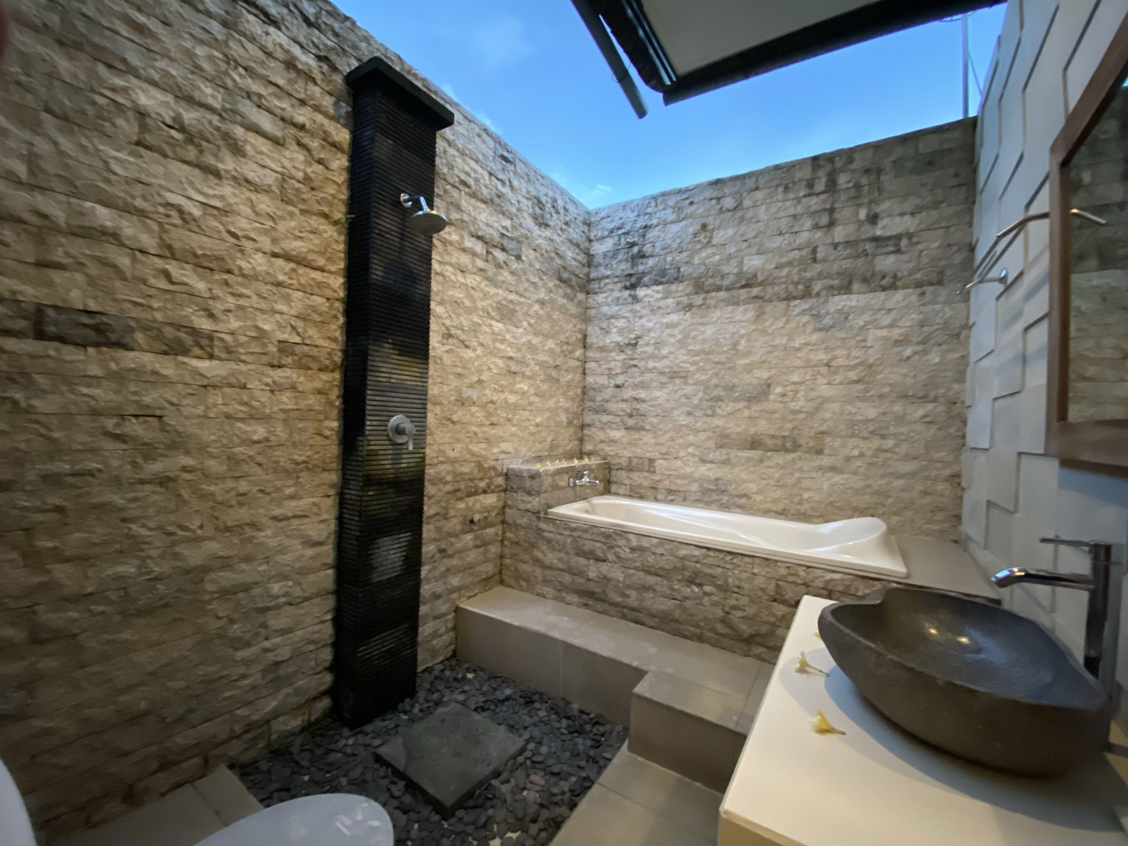 Un baño con pared de piedra, un elemento vertical negro, una bañera blanca y un lavabo grande sobre el mostrador.