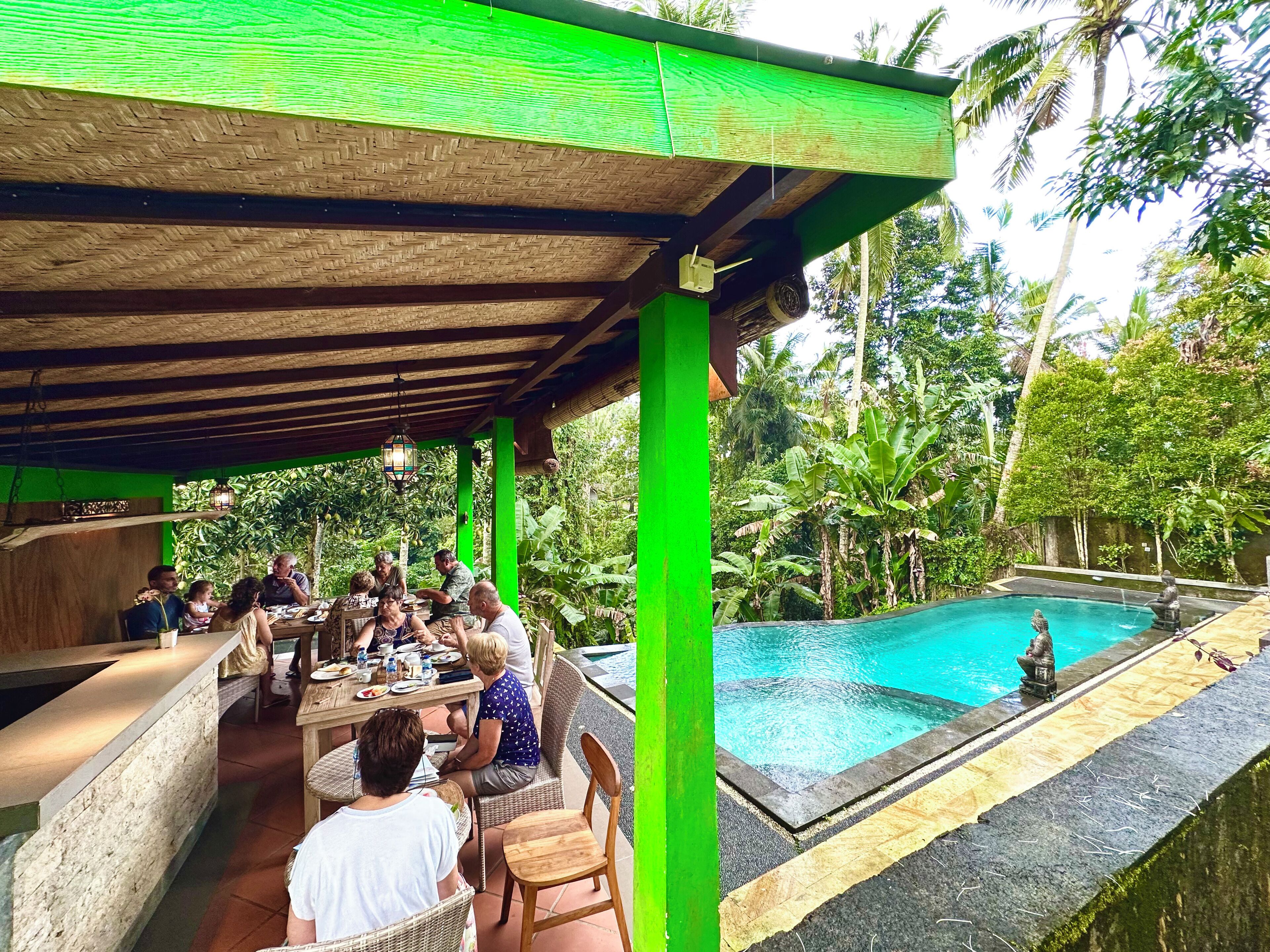 Uma área de refeições ao ar livre coberta, com piscina e vegetação exuberante ao fundo.