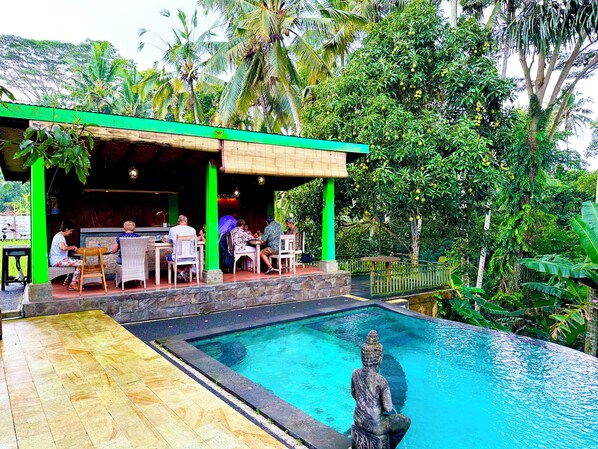 A poolside area with a Buddha statue, dining tables, and chairs, surrounded by lush greenery and palm trees. - Dupa Ubud Villa (Ubud)