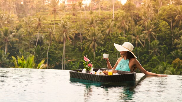 Una persona relajándose en una plataforma flotante en una piscina, con un paisaje tropical de fondo.
