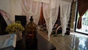 Una habitación con una cama, una mesita con una botella y una planta, y una ventana con cortinas.