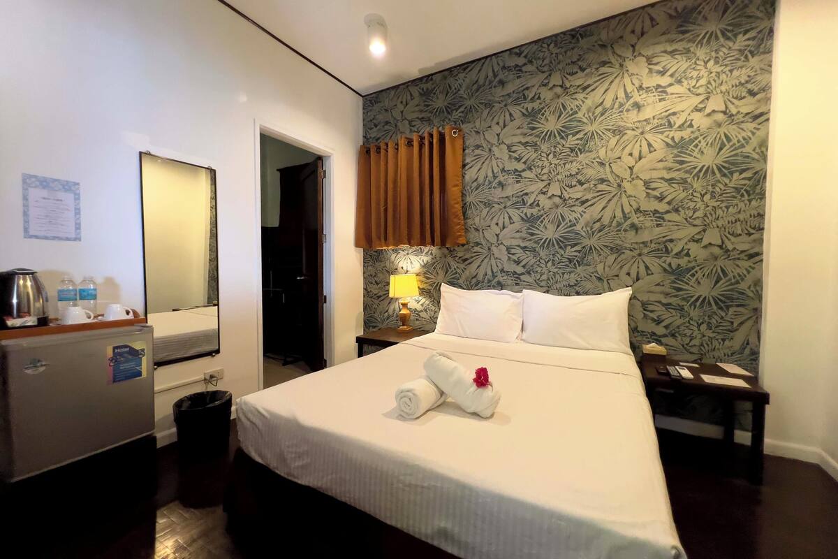 Una habitación de hotel con una cama, una mesita de noche, una lámpara, un espejo, un escritorio con hervidor y un cubo de basura.