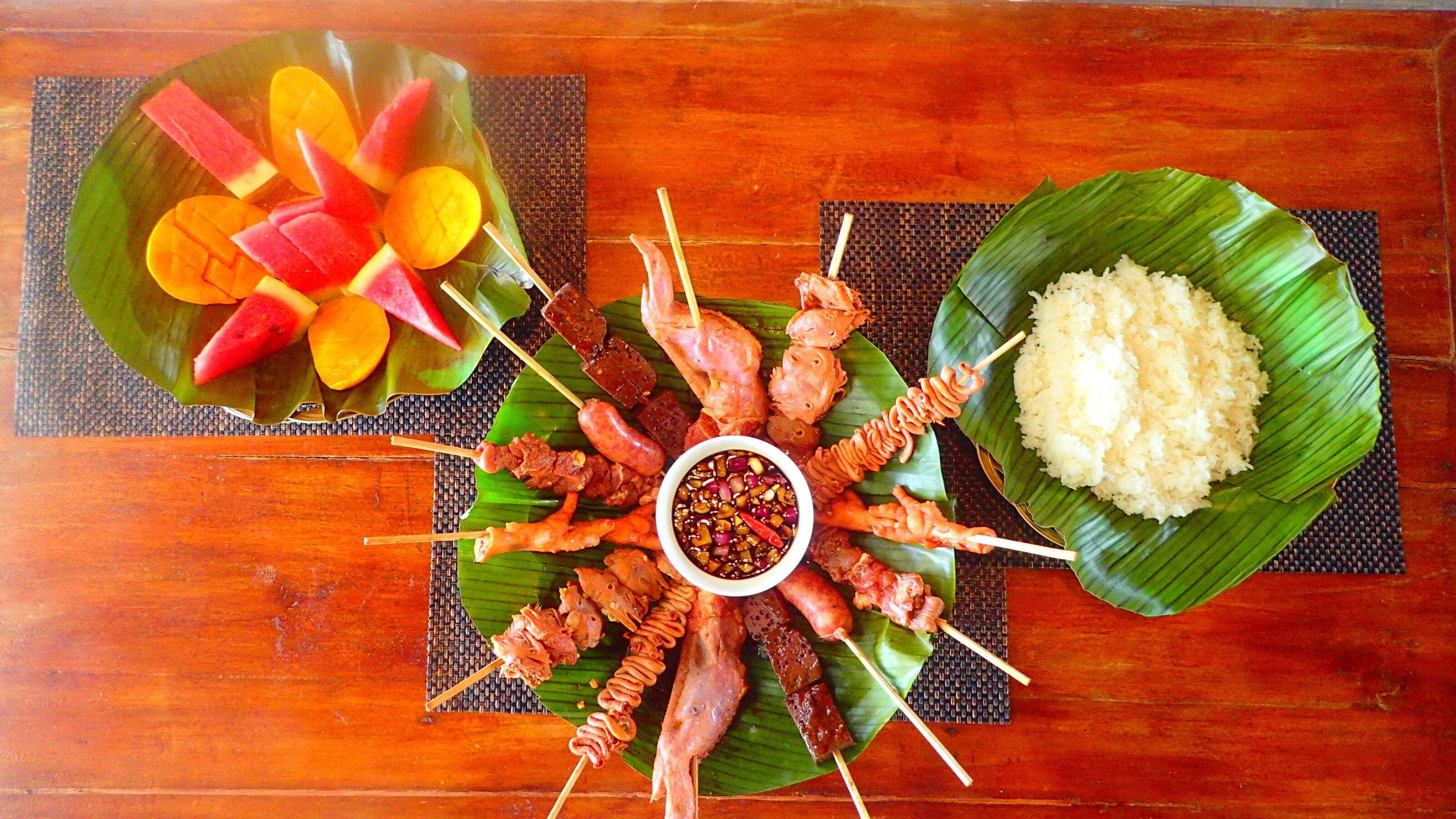 A wooden table with a variety of skewered meats, a bowl of rice, and a bowl of fruit.