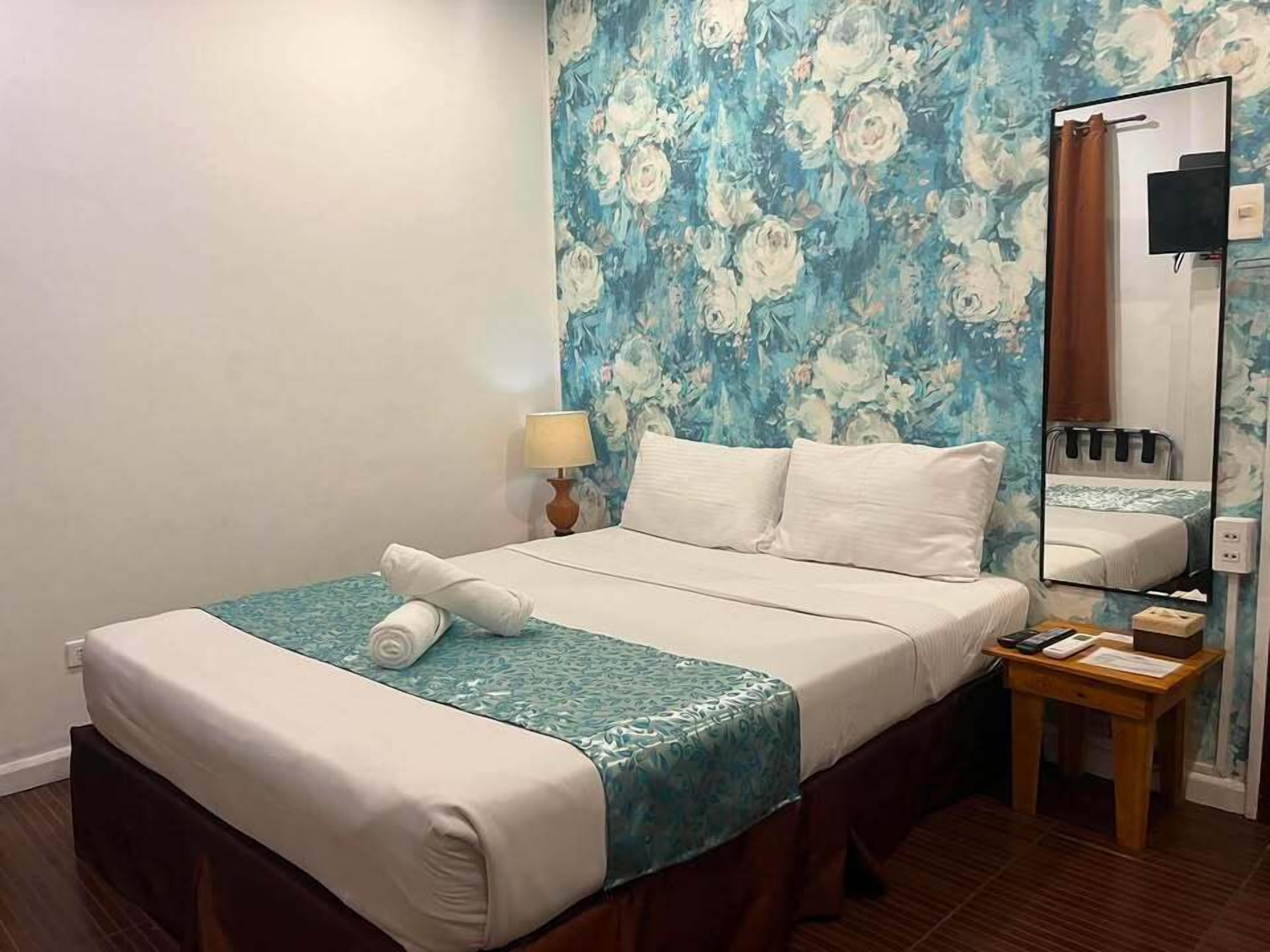 Una habitación de hotel con cama, mesita de noche, lámpara, espejo y papel tapiz con motivos florales.