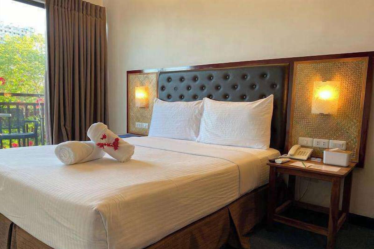 Una cama bien hecha con ropa blanca, un cojín decorativo y una mesita de noche con un teléfono y una lámpara.