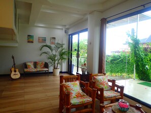 A spacious living room with a wooden floor, a sofa, two bamboo chairs, a guitar, and a large window offering a view of a garden. - Aozora Seaside Mactan (Lapu-Lapu)