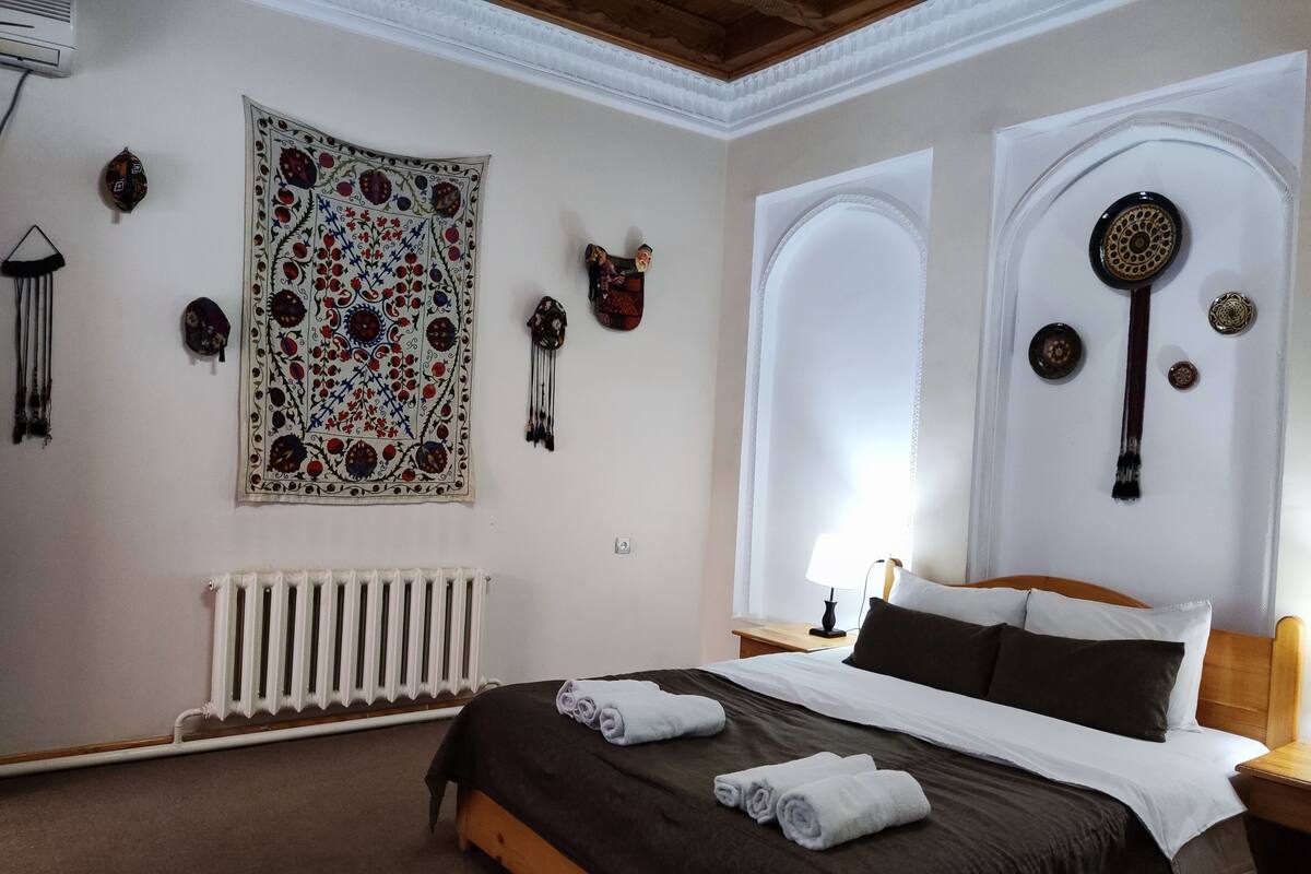 Quarto com uma cama grande, um radiador, um tapeçário na parede e uma janela com detalhes decorativos.
