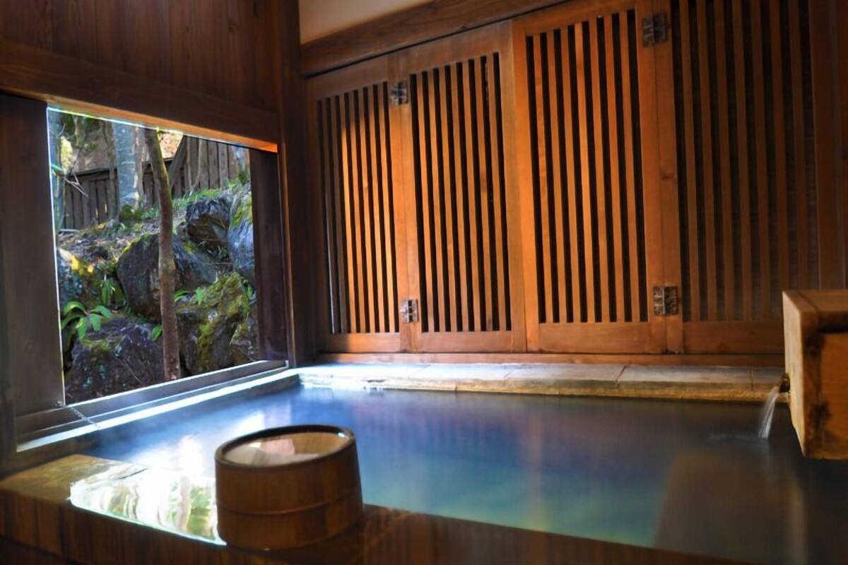 Un onsen japonais traditionnel, avec un intérieur en bois, une vue sur une zone boisée et un tonneau en bois placé au bord de la source thermale.