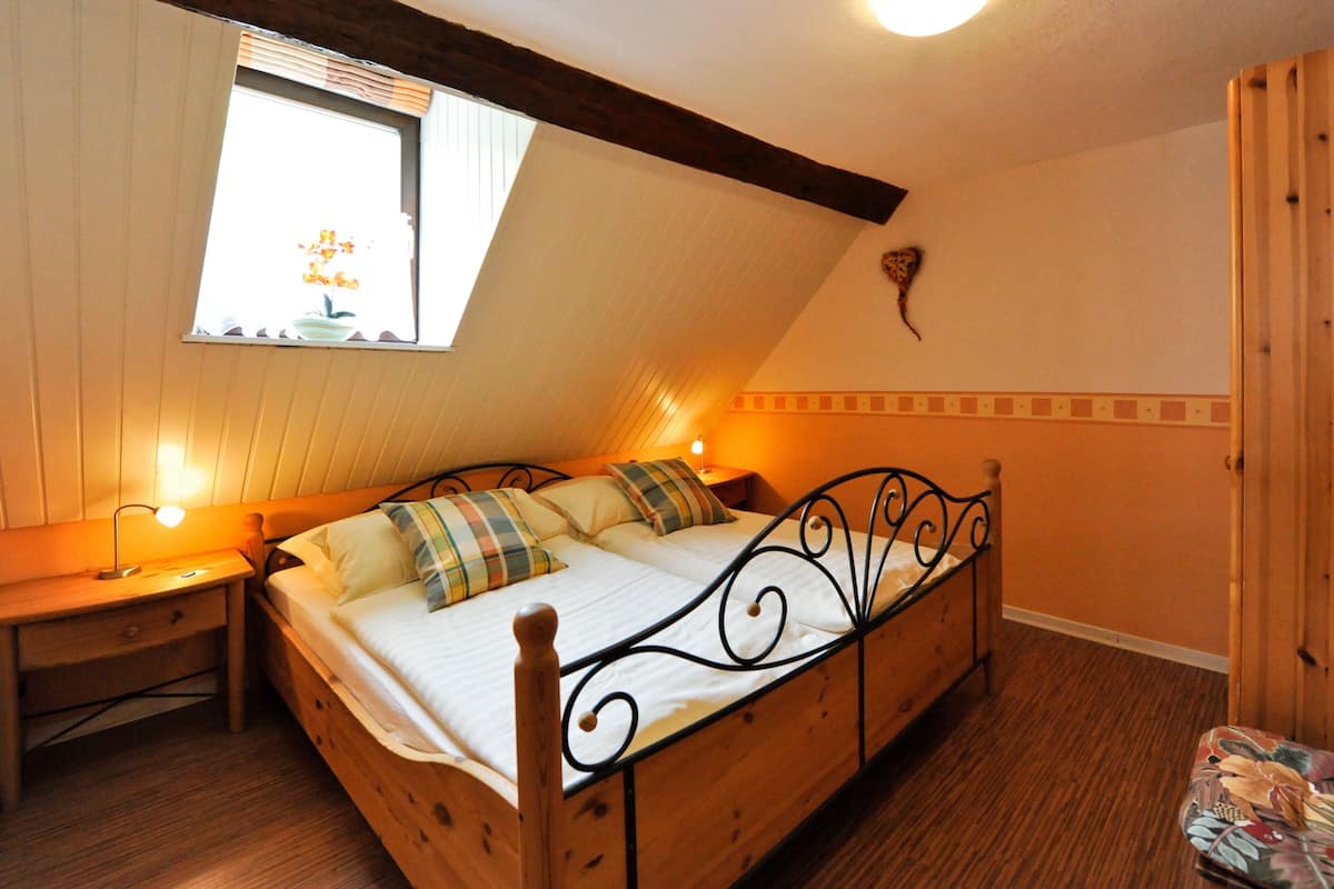 Ein Einzelbett mit Holzbettgestell und Kopfteil, ein Nachttisch mit Lampe, ein Fenster mit Blick auf Blumen und ein Holzschrank.