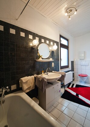 Une salle de bain moderne avec une baignoire, un lavabo et un miroir. Une fenêtre laisse entrer la lumière naturelle et un tapis rouge recouvre le sol.