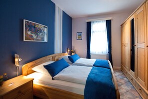 Un lit double avec une literie bleue, une table de chevet avec une lampe, un tableau au mur et une fenêtre avec des rideaux.