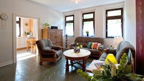 Una sala de estar con sofá, sillones, una mesa de centro y una zona de comedor con mesa y sillas. Hay ventanas con cortinas, un reloj en la pared y una lámpara sobre la mesa.
