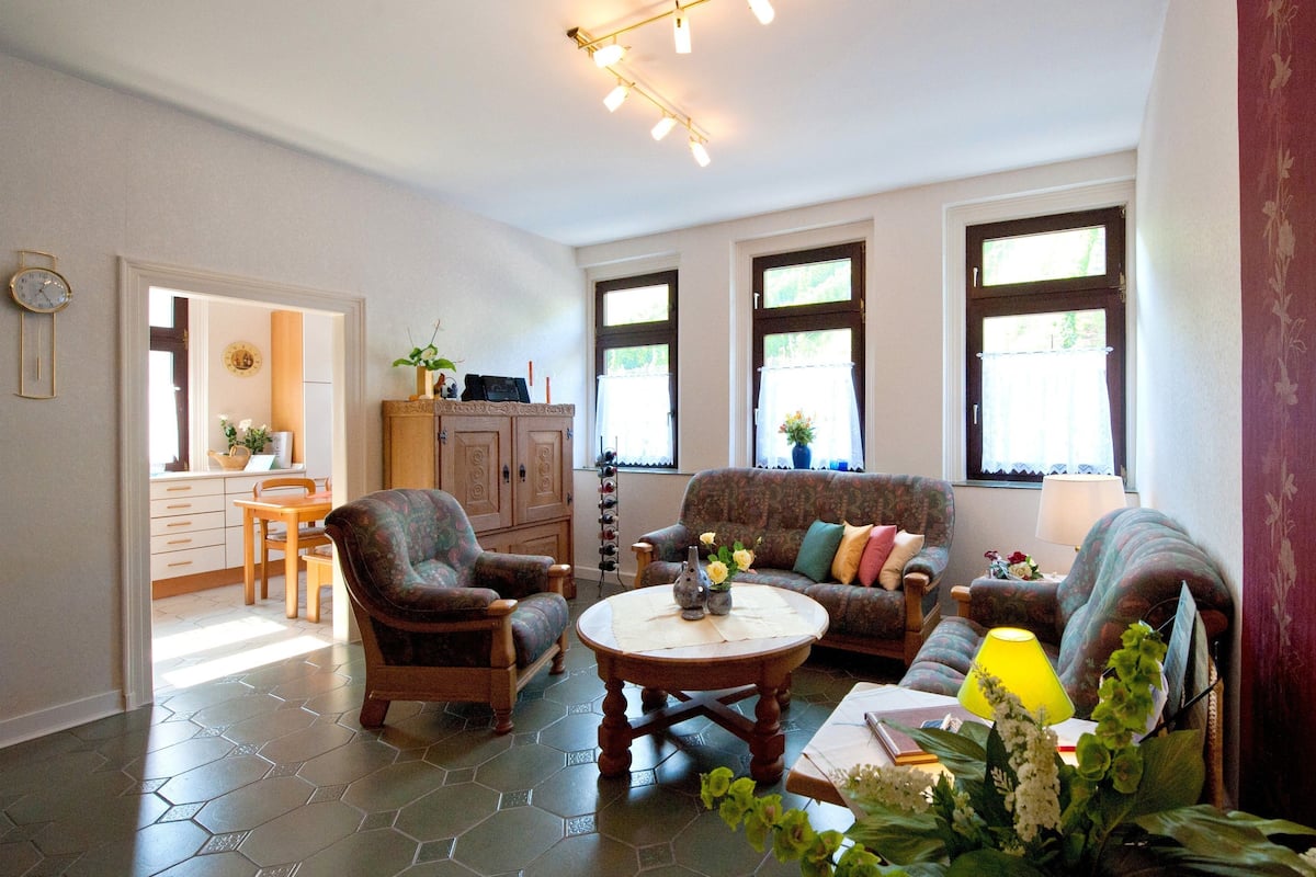Ein Wohnzimmer mit einer Couch, Sesseln, einem Couchtisch und einem Essbereich mit Tisch und Stühlen. Es gibt Fenster mit Vorhängen, eine Uhr an der Wand und eine Lampe auf dem Tisch.