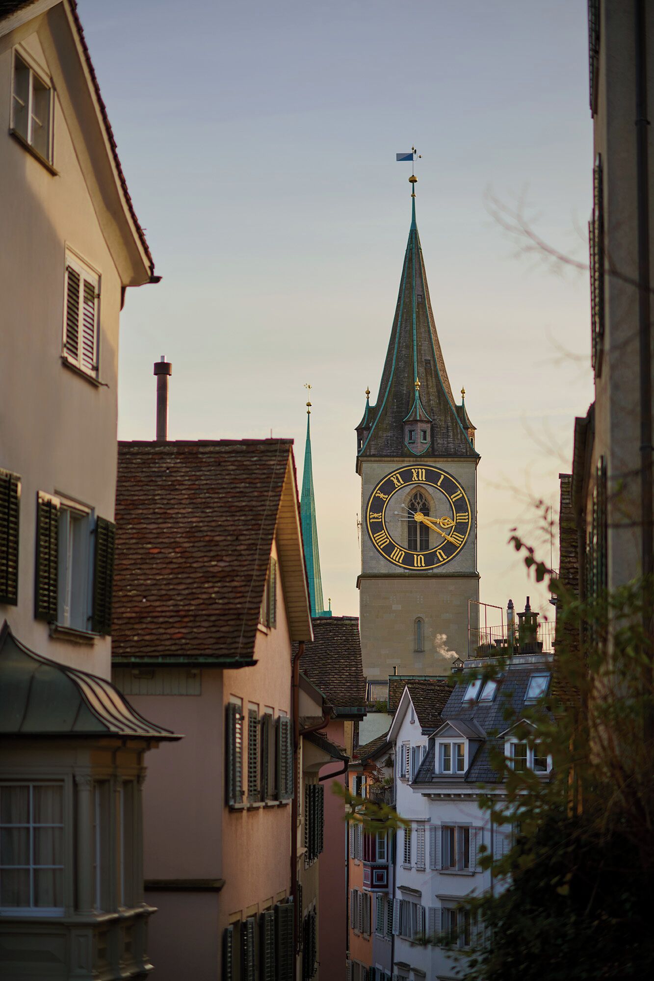 Ein historischer Glockenturm mit spitzem Dach und Wetterfahne, umgeben von traditionellen europäischen Gebäuden mit verriegelten Fenstern.