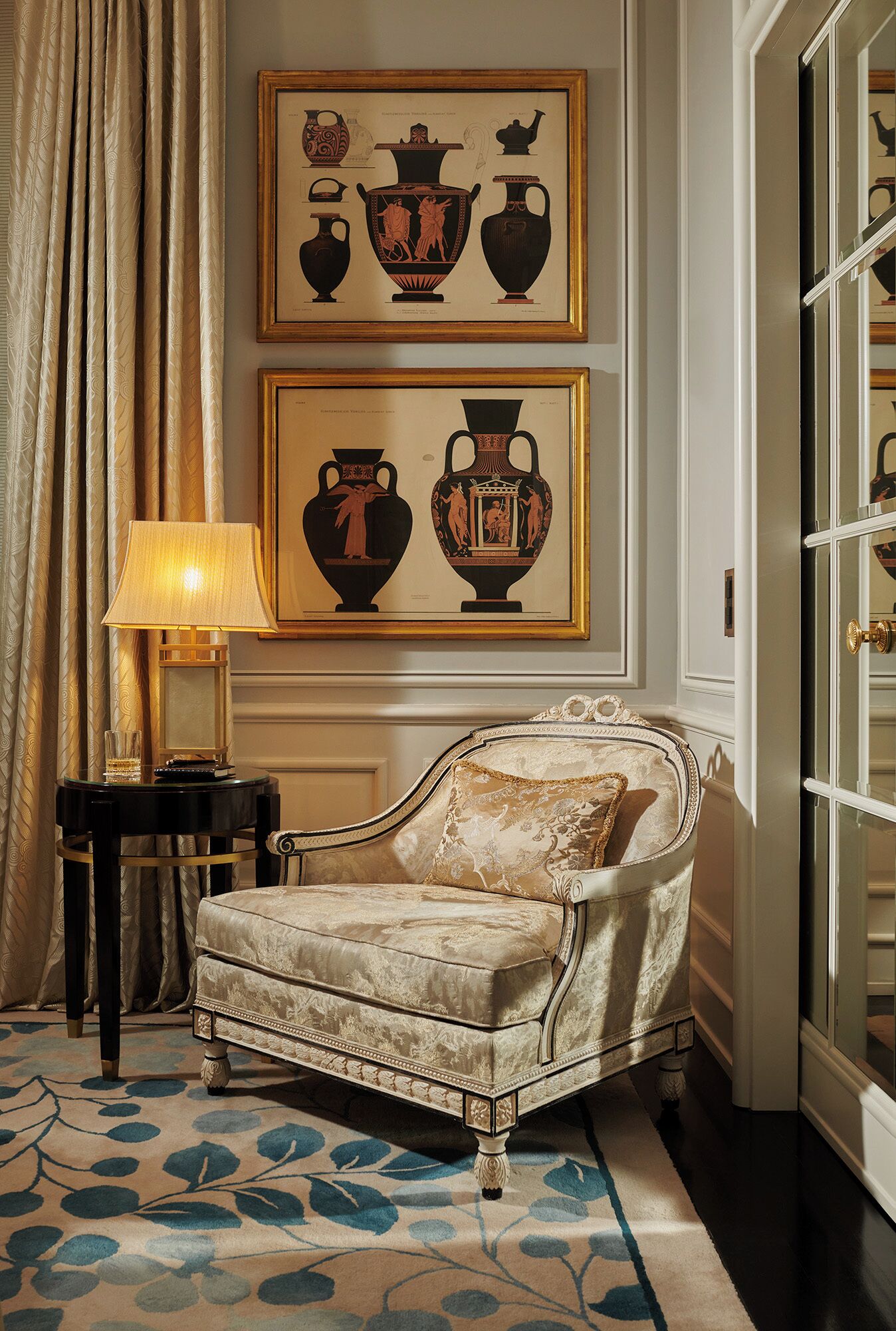 A room with a classic armchair, a framed artwork of vases, a lamp, and a patterned carpet.