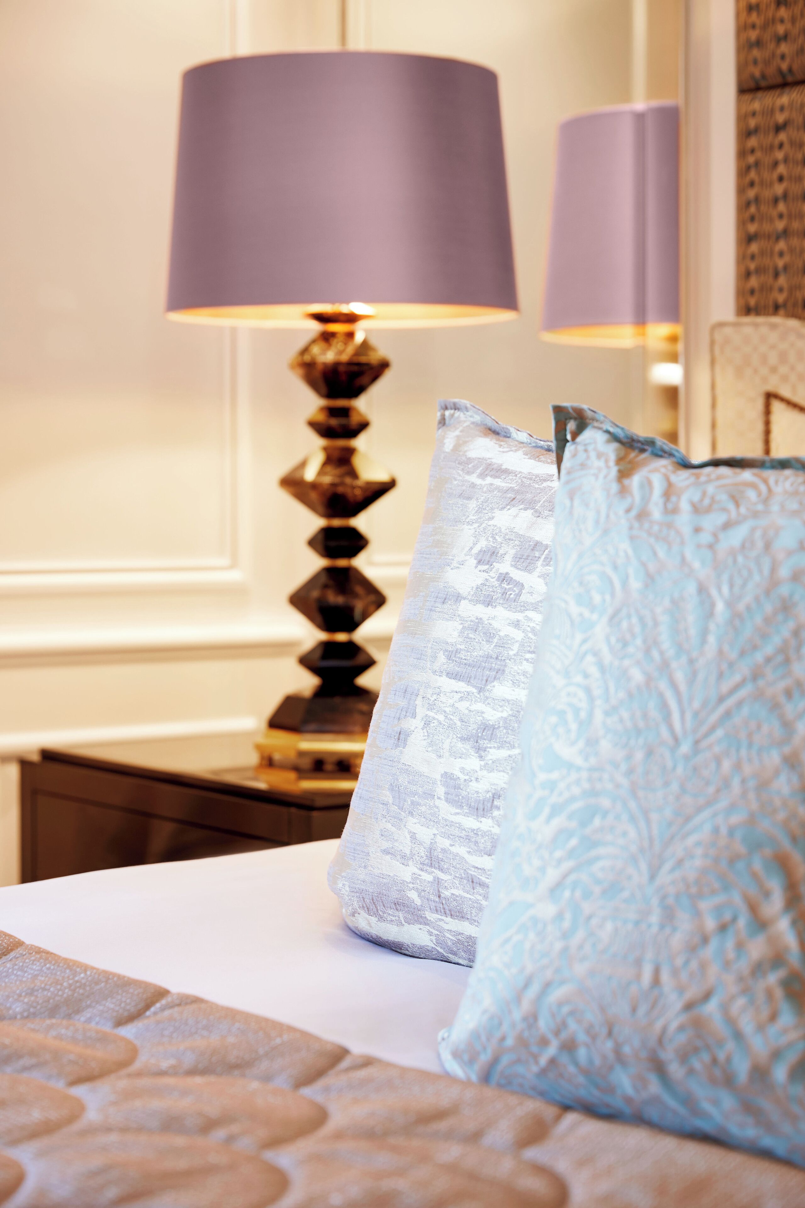 A bed with patterned bedding and two pillows, a lamp with a purple shade, and a patterned wall in the background.