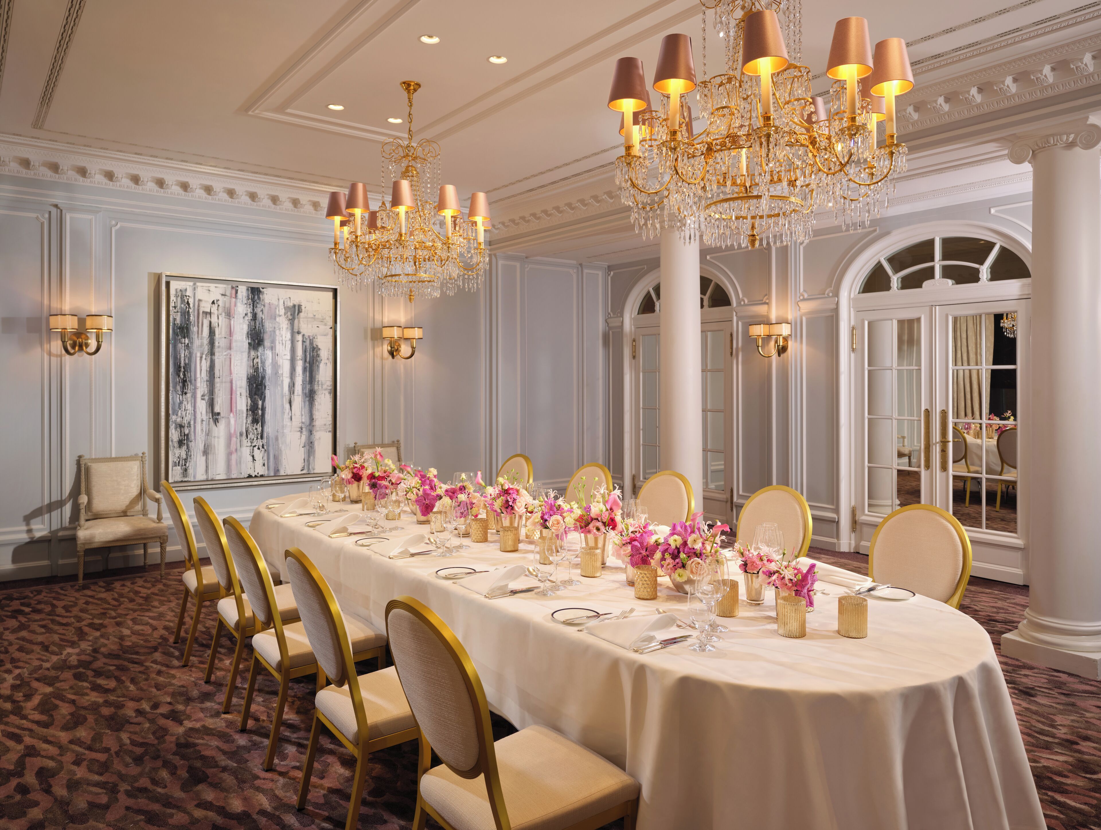 A formal dining room with a long table set for a meal, surrounded by chairs, and adorned with floral centerpieces.