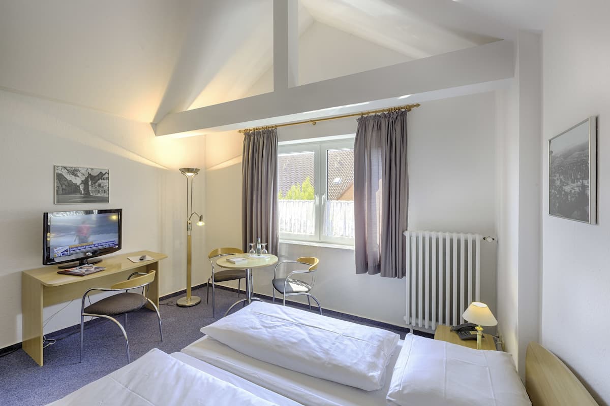 A hotel room with two beds, a desk with a TV, a chair, a small table, and a window with curtains.