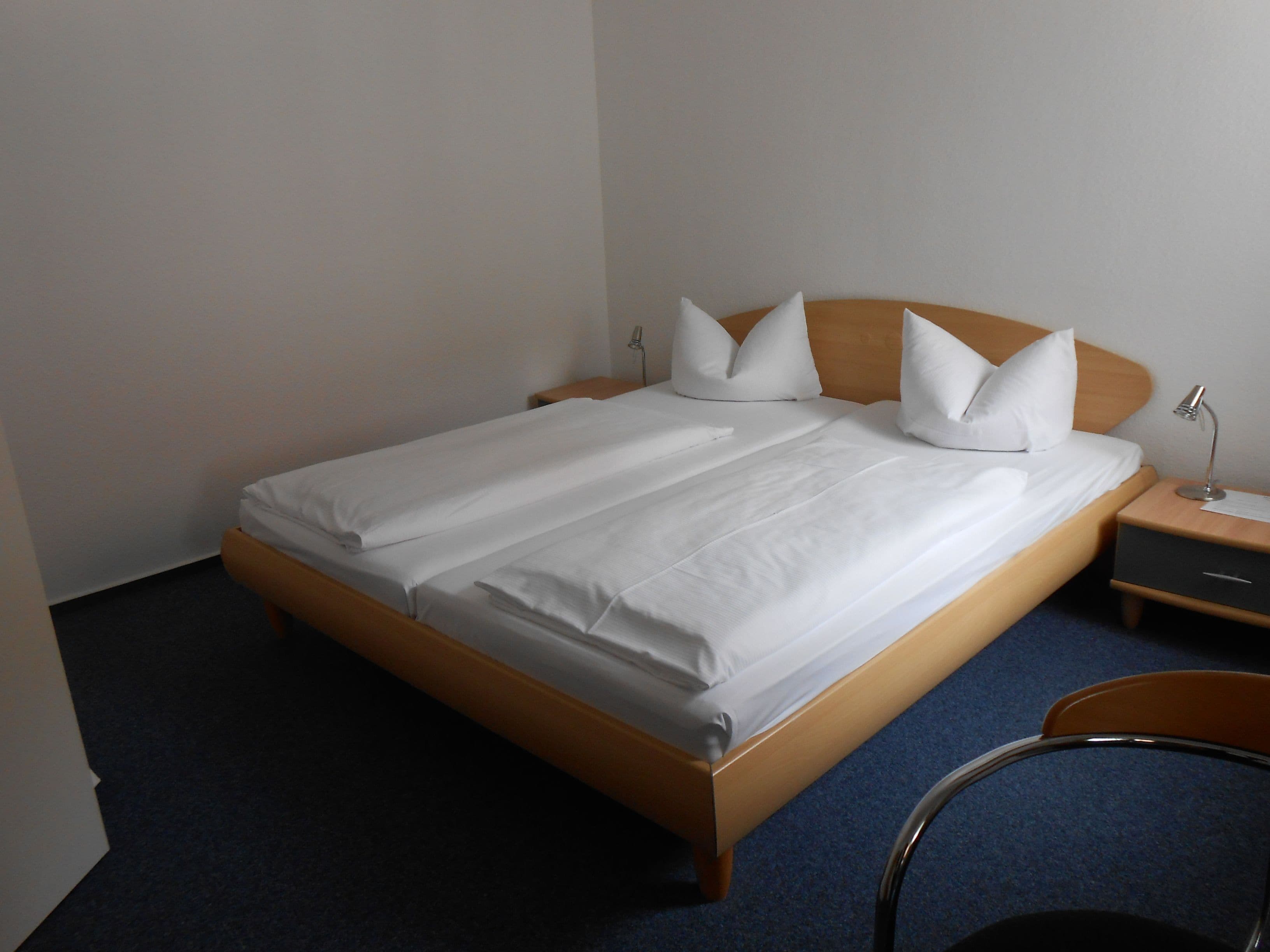 A neatly made bed with white linens and pillows, a wooden headboard, a bedside table with a lamp, and a chair.