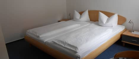 Una cama bien tendida con sábanas y almohadas blancas, un cabecero de madera, una mesita de noche con una lámpara y una silla.