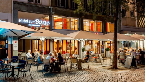 An outdoor seating area of a restaurant with tables and chairs, umbrellas, and a building with a sign reading "Hotel zur Borse". - Hotel zur Börse (Hamelin)