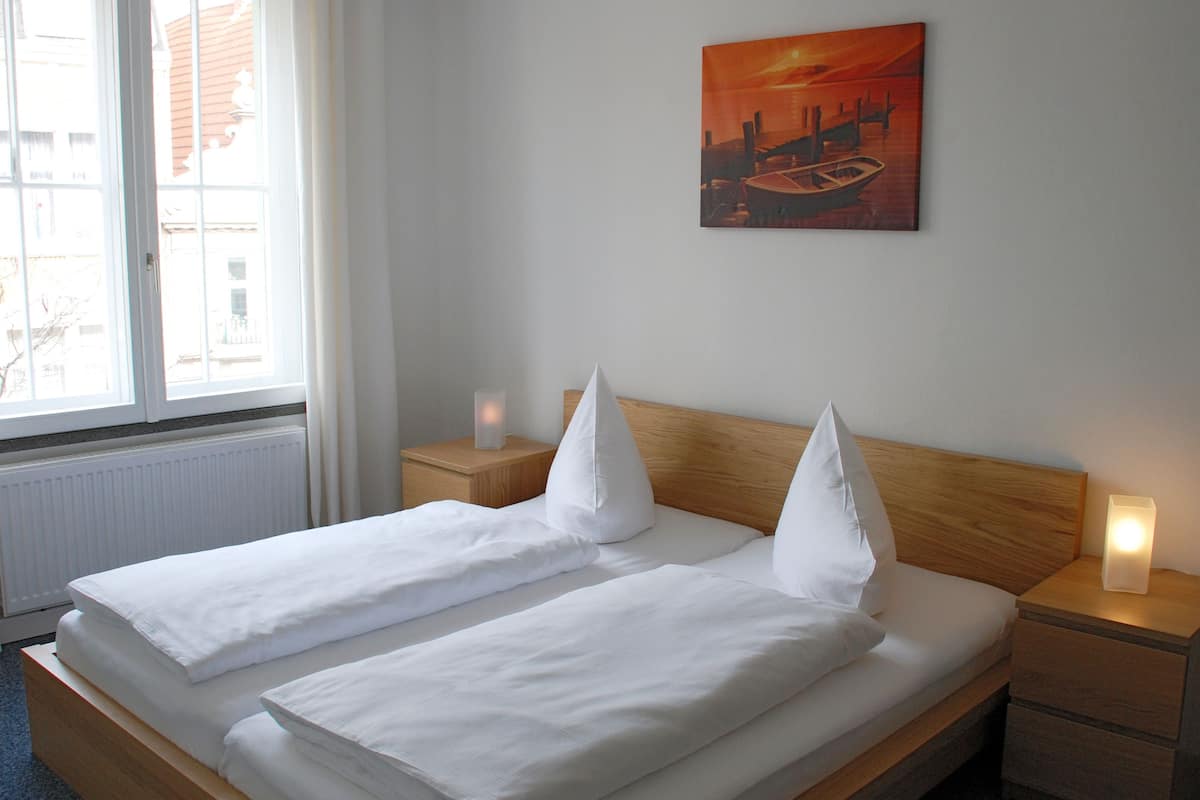 A neatly made double bed with white linens and pillows, a wooden headboard, bedside tables with candles, and a painting of a sunset on the wall.