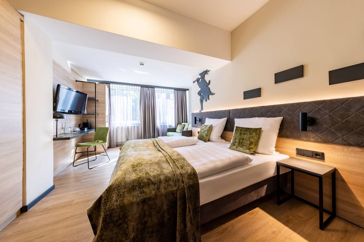 A modern hotel room with a large bed, a desk with a flat-screen TV, a chair, and a window with curtains.