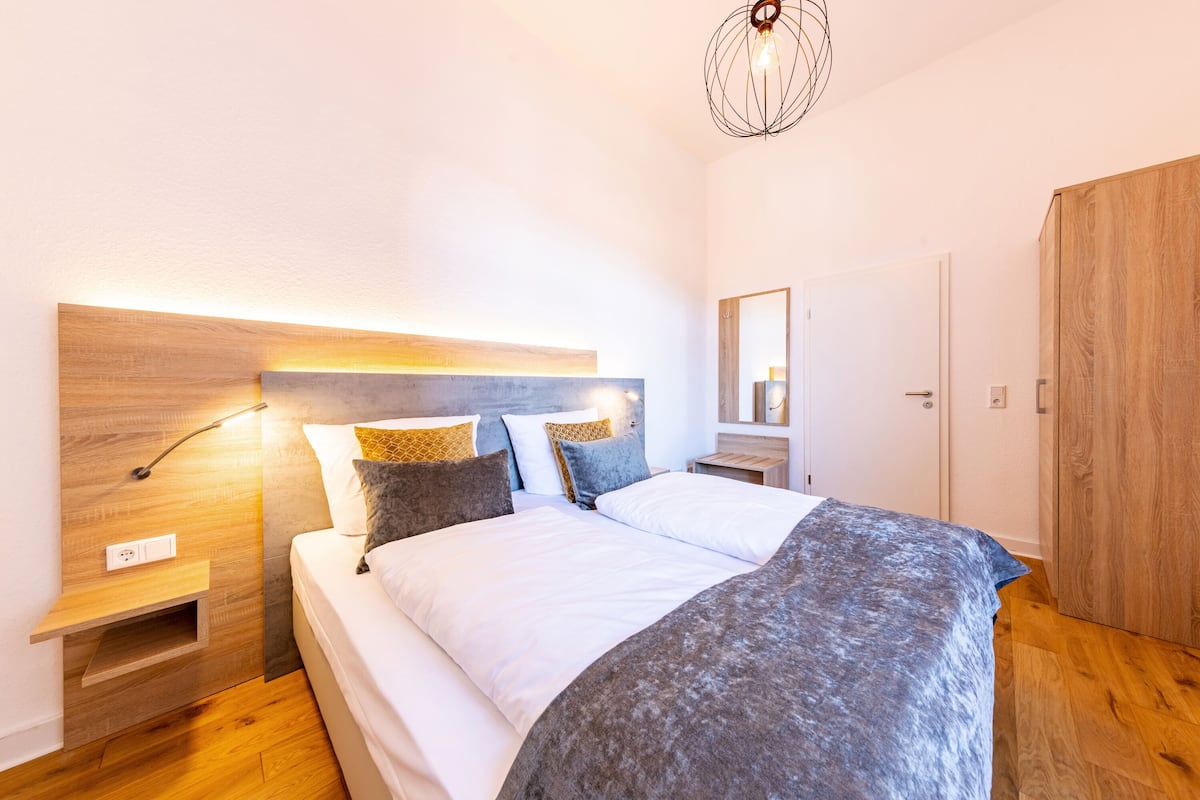A modern bedroom with a wooden headboard, a bedside table with a lamp, and a large bed with a grey blanket.