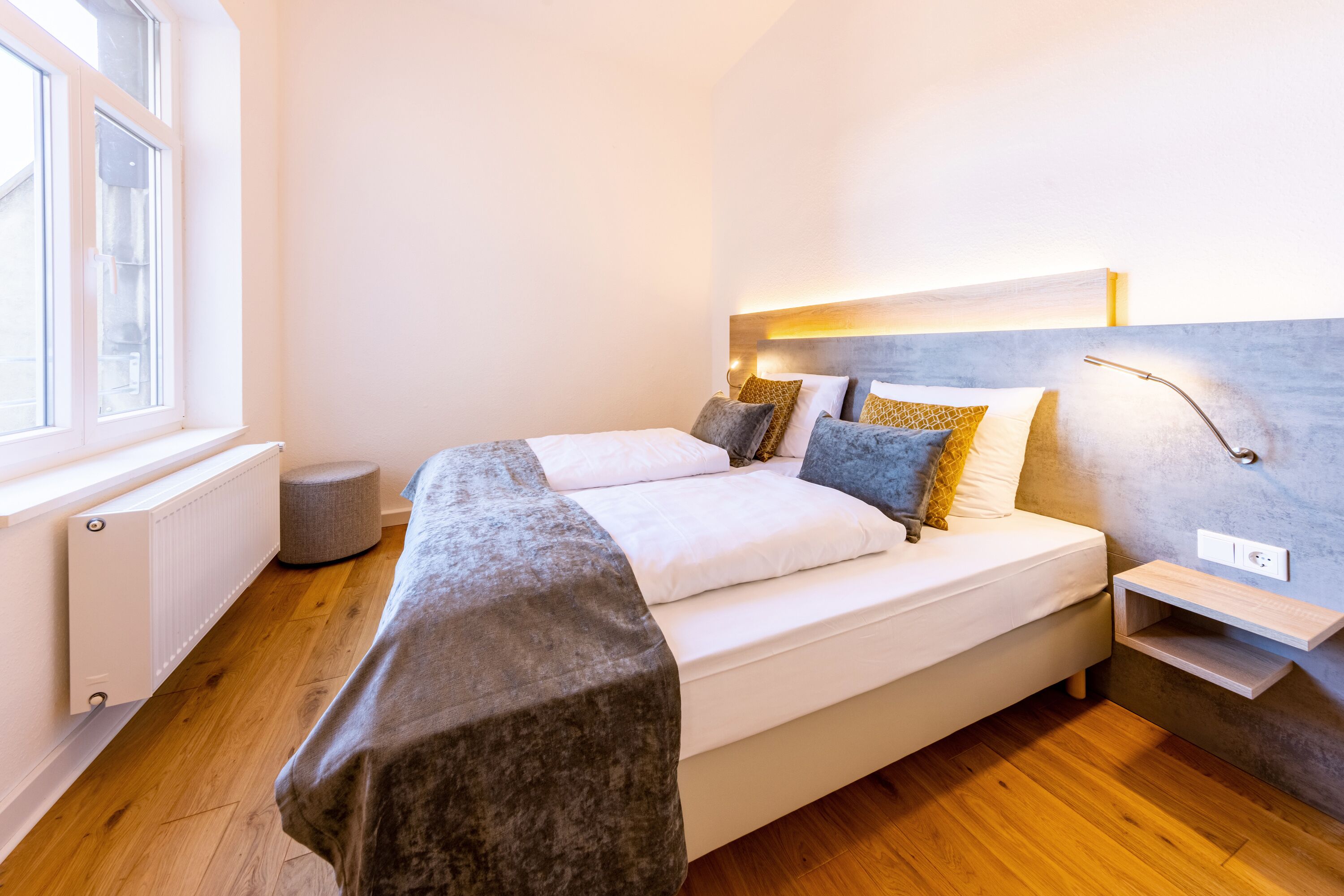 A modern bedroom with a large bed, bedside tables, a round ottoman, and a window.