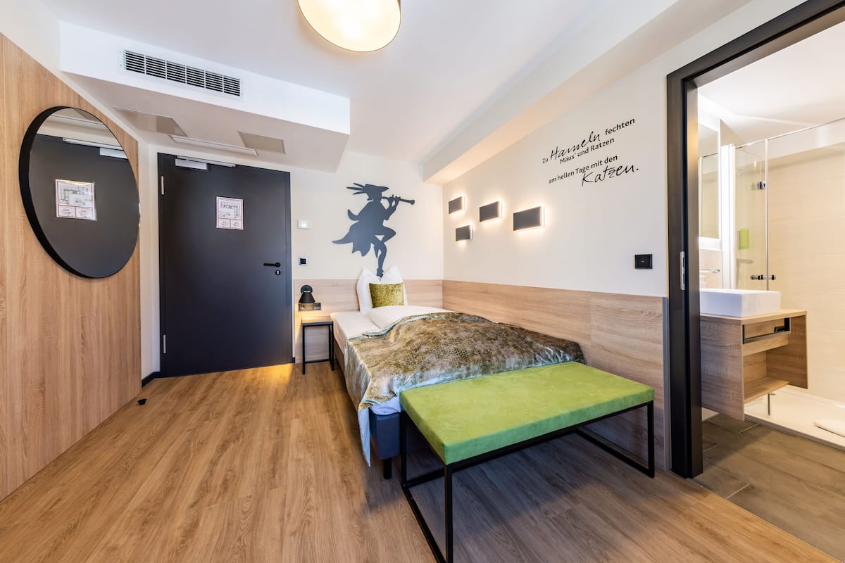 A modern hotel room with a bed, bedside table, and a green bench. There is a wall with a silhouette decoration and a motivational quote.