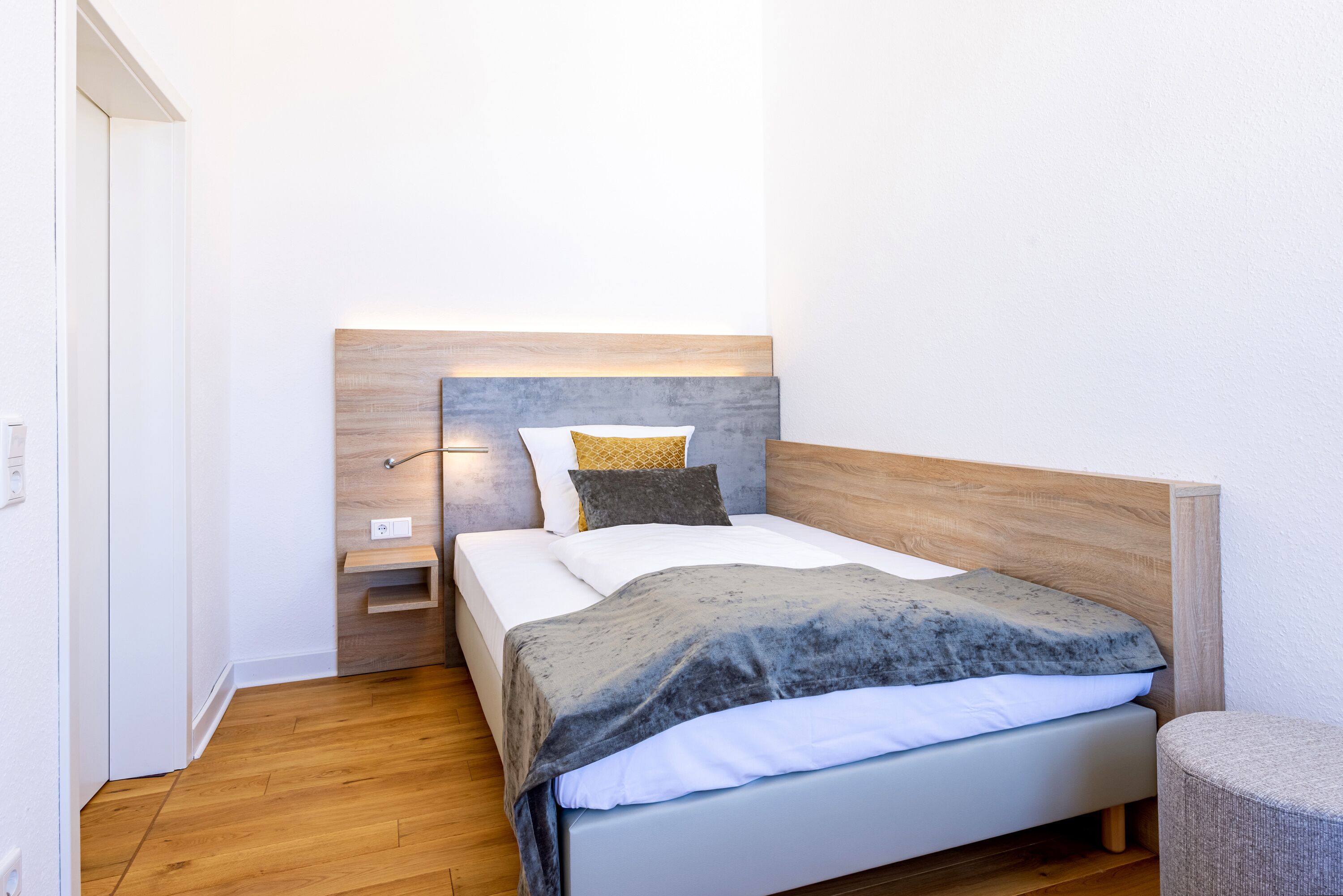 Ein ordentlich bezogenes Bett mit Holz-Kopfteil, Nachttisch und Lampe.