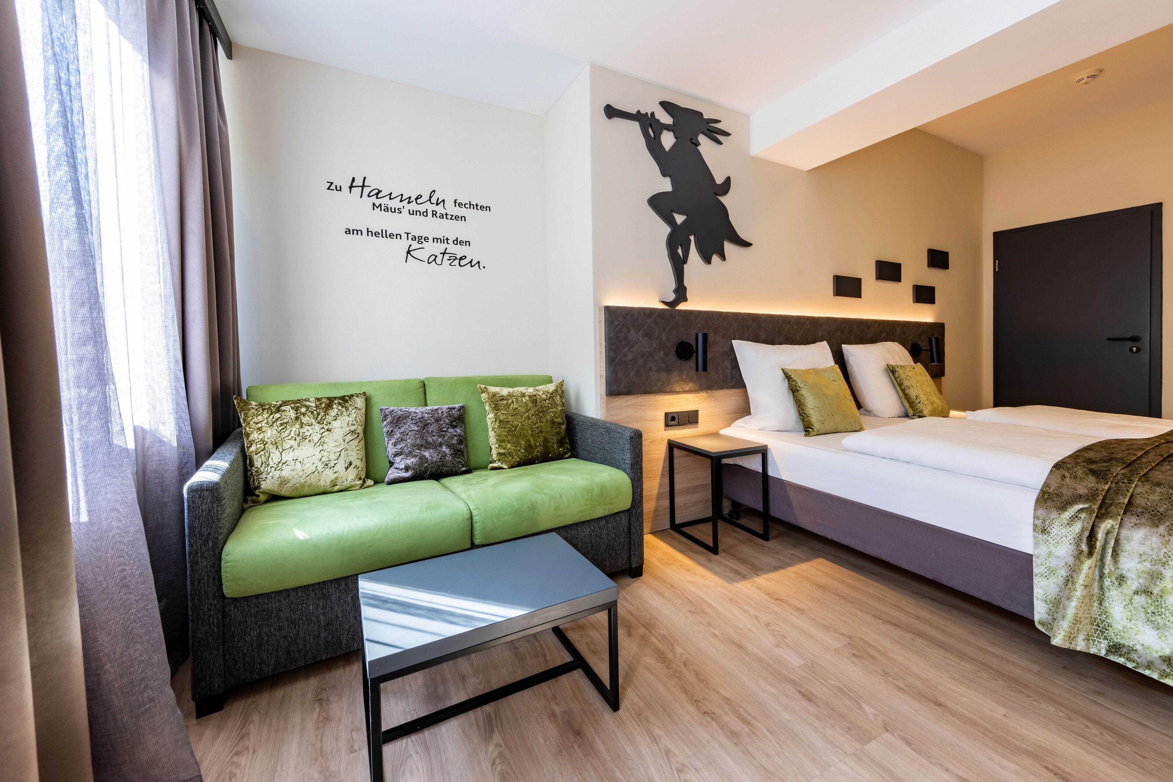 A modern hotel room with a green sofa, a small table, a bed with white bedding, and a wall with a silhouette decoration.