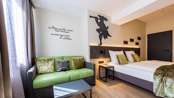 A modern hotel room with a green sofa, a small table, a bed with white bedding, and a wall with a silhouette decoration.